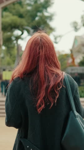 Vertical video, A young woman with long, red hair walks through a park. She's wearing a dark coat and carrying a black bag. The park has trees and steps leading up to a building.