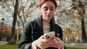 A young woman with red hair is using her smartphone and wearing earbuds in a park. She looks focused and is texting. - Powered by Shutterstock - Get 15% off with code: PIKWIZARD15