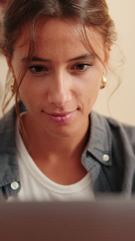 Vertical video, Young woman with brown hair, working on laptop, chatting on social networks sitting at table in living room, raises head and smiles looking at camera