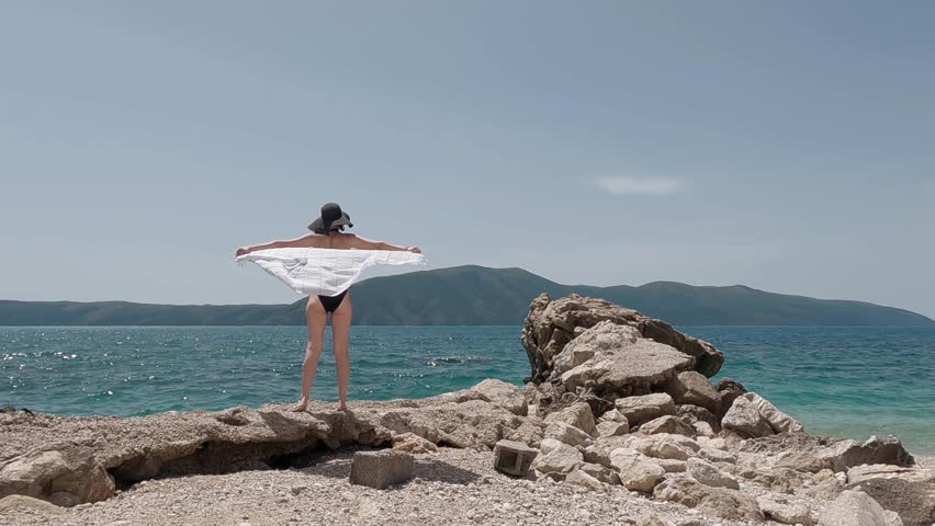 Stylish woman in a wide-brimmed black hat holds a flowing white fabric against the summer sky, standing on a rocky beach in Vlore, Albania