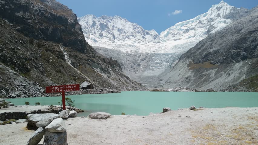 A first-person point-of-view shot shows a tourist hiking past the stunning, turquoise glacial waters of Laguna Llaca at the base of snow-capped peaks in Peru's Huascarán National Park.