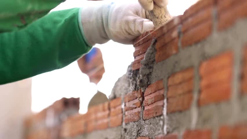 A skilled worker is laying bricks on a construction site. He is focused on applying mortar with a trowel, showcasing craftsmanship in masonry work.
