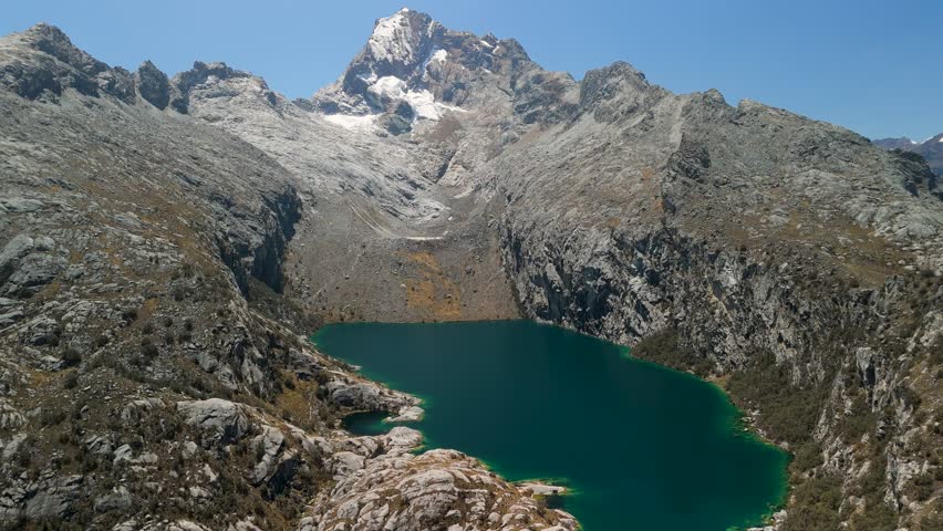 Drone pulls back to reveal stunning turquoise Laguna Churup surrounded by rugged Andes mountains under a clear blue sky in Peru.