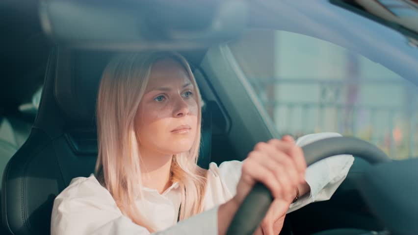 Exhausted blonde woman touching forehead while gripping steering wheel tightly. Closing eyes and leaning back after stressful moment. Processing frustration or feeling mentally drained inside car.