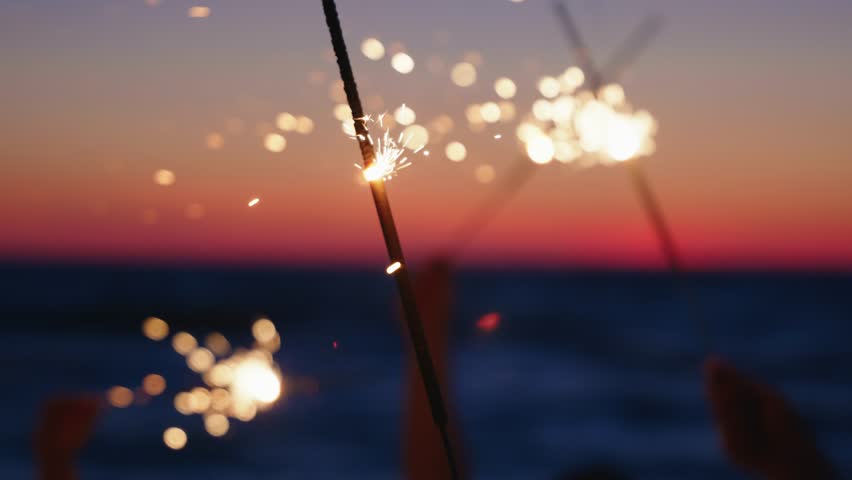 Glowing sparklers held against a colorful sunset sky, creating a joyful ambiance by the ocean, perfect for celebrations and gatherings