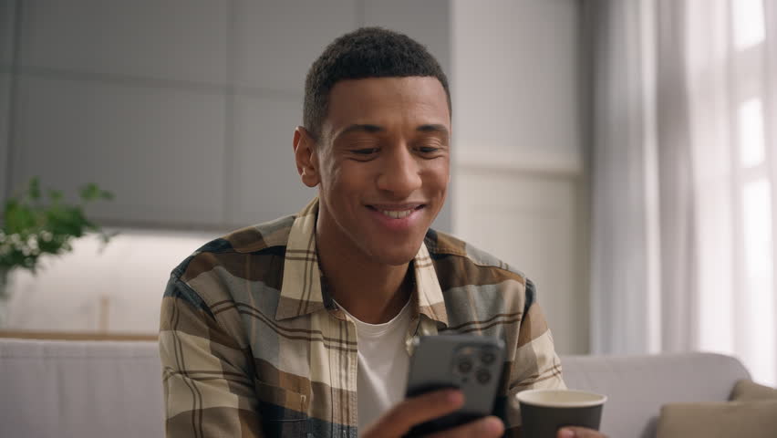 Cheerful satisfied smiling African American man guy male browsing smartphone drinking tea coffee scrolling social media application resting at couch home using mobile phone gadget addict user chatting - Powered by Shutterstock - Get 15% off with code: PIKWIZARD15