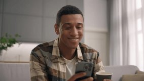 Cheerful satisfied smiling African American man guy male browsing smartphone drinking tea coffee scrolling social media application resting at couch home using mobile phone gadget addict user chatting - Powered by Shutterstock - Get 15% off with code: PIKWIZARD15