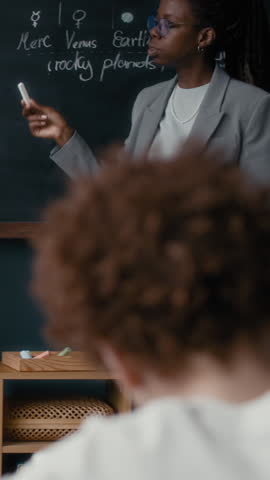 Vertical of African American teacher discussing solar system at blackboard as students participate, one drawing planets and another raising hand