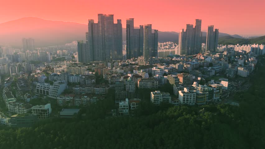 Aerial view of a sprawling modern cityscape featuring towering skyscrapers, residential buildings, and lush green forests, illuminated by a vibrant sunset sky in South Korea.