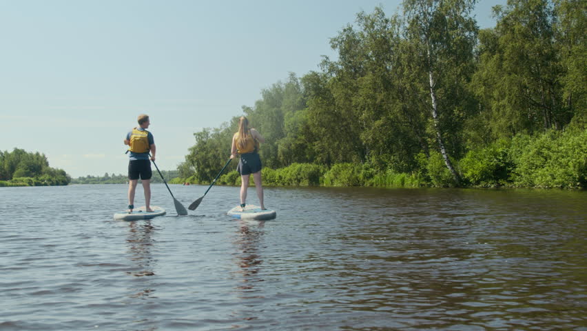 Wide back view shot of young couple in life jackets moving across river using paddleboards during outdoor training