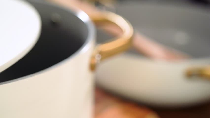 A close-up shot of a white ceramic pot with a gold handle shows the lid resting on the rim. The composition emphasizes minimal design and soft lighting.