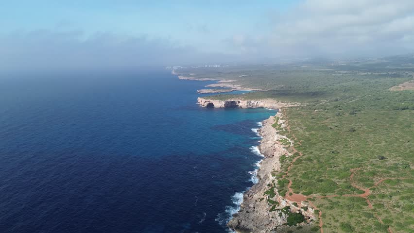 Wide 4K drone shot of Mallorca’s coastline featuring the blue sea on the left and lush green hills on the right, under a sunny sky with scattered low clouds. Captured June 2025.