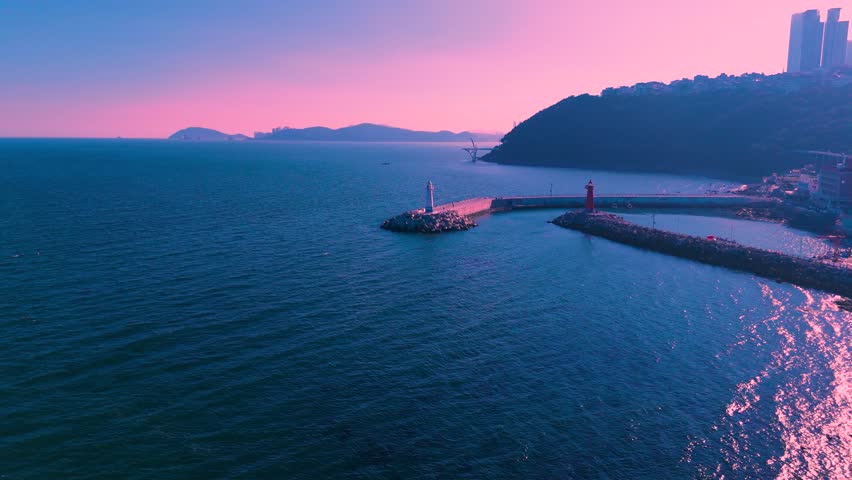 Aerial view of two lighthouses on breakwaters extending into an ocean, with a coastal city built on a hill under a pink and purple sky, Busan, South Korea.
