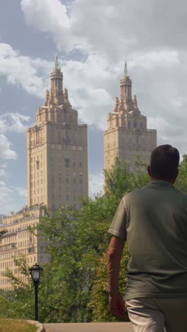 A vertical view of a man walking along a quiet path in Central Park, New York City, with the iconic twin towers of an upscale apartment building rising in the background behind lush green trees.