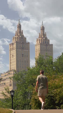 A vertical view of a man walking along a quiet path in Central Park, New York City, with the iconic twin towers of an upscale apartment building rising in the background behind lush green trees.