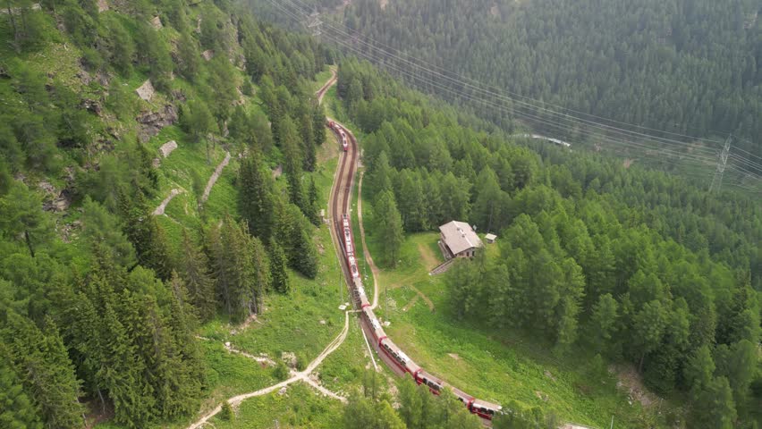 Top-down drone view of a red train snaking through dense conifer forest on a steep alpine slope with trails and a small cabin.