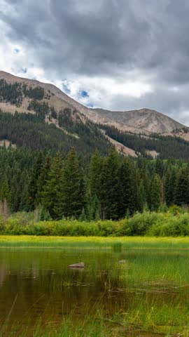 Vertical Timelapse. Mountain Landscape, Conifer Forest, Alpine Lake and Clouds Above Peaks