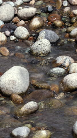 Close Up Of River Water Flowing Over Smooth Stones. Camera Pans From Right To Left, Revealing A More Turbulent And Rapid Current. Natural Waterway Dynamics. Vertical