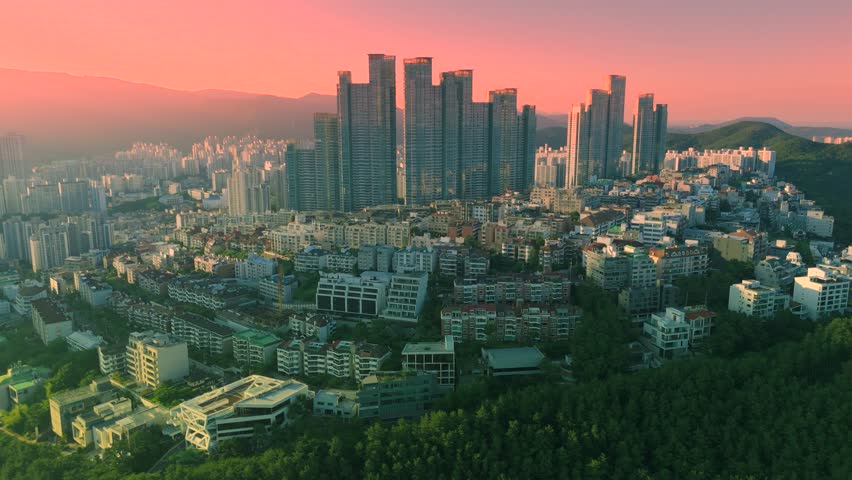 Aerial view of a dense urban cityscape with skyscrapers, residential buildings, green hills, and trees under an orange and pink sky, Busan, South Korea.