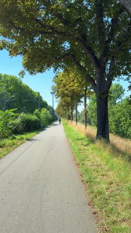 Cyclist on sunny tree lined path heading towards telecommunications tower in Mannheim, Germany, vertical video
