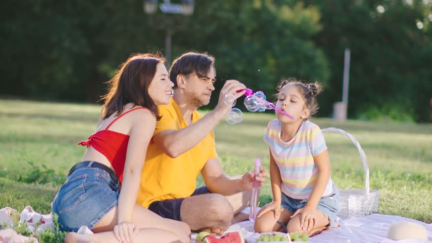 Family enjoys a sunny afternoon in the park, making bubbles and having a picnic with fresh fruits and laughter in a green grassy area