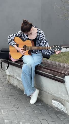 Young Caucasian woman playing guitar and singing while sitting on a bench. 
