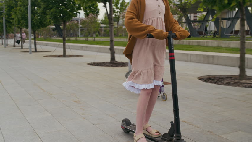 Two active youthful girls in casual attire riding on skateboard and electric scooter along sidewalk with row of green trees and lawns surrounded by concrete borders