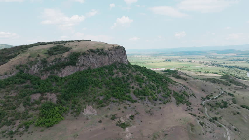 A super wide aerial drone shot makes a sweeping orbit around the majestic Măgura Uroiului mountain in Romania, revealing the sheer rock cliff and the vast surrounding plain.