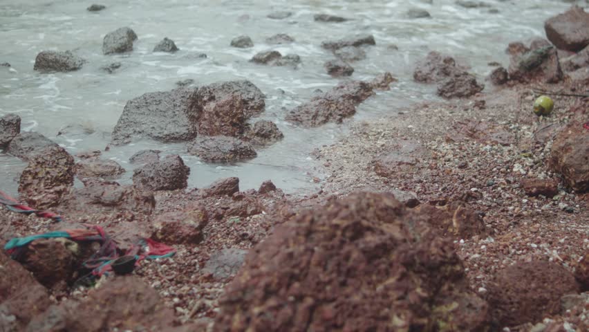 Detailed view of Goa’s shoreline with reddish laterite rocks and broken shells scattered across wet sand, as gentle waves approach under a cloudy sky.