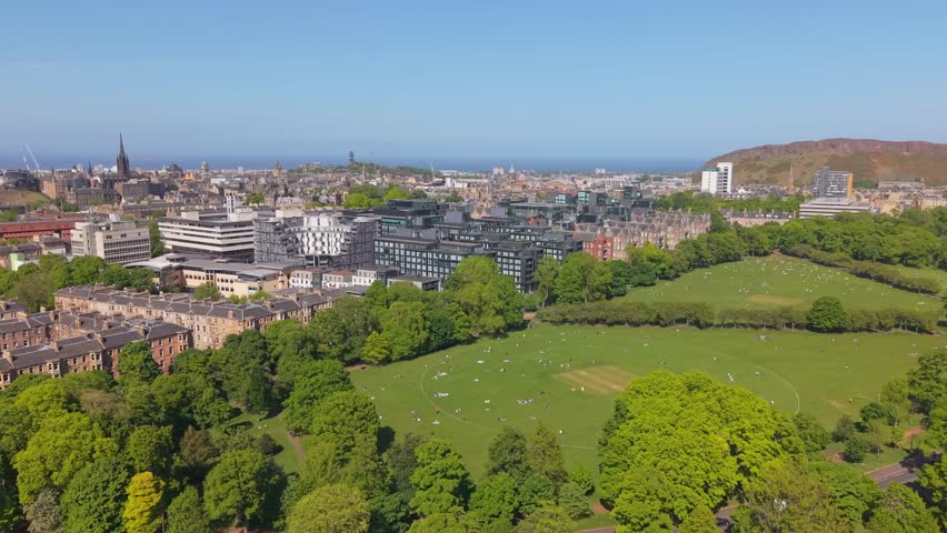 Drone flies over green Edinburgh meadows, cityscape to the left, and expansive Holyrood Park stretching in the background under clear skies