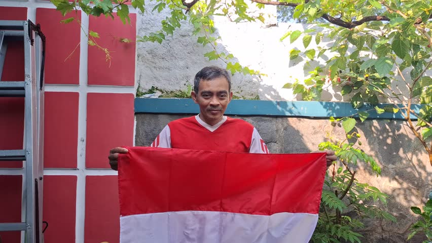 An Indonesian man wearing a red shirt is waving the red and white flag which is the flag of the unitary state of the Republic of Indonesia (NKRI). this man is celebrating Indonesian Independce day.