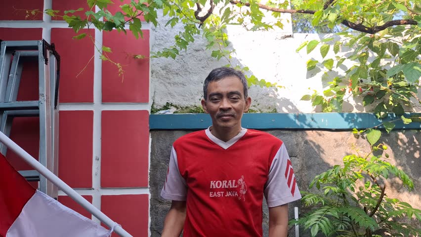 A man waving the red and white flag, the flag of the Unitary State of the Republic of Indonesia, in front yard.  The camera slowly zooms in on him. Celebrate Indonesian Independence Day on August 17th