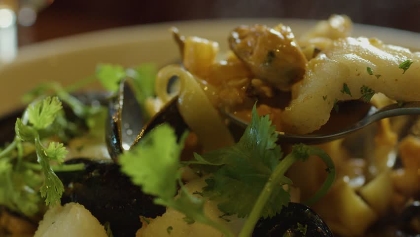 A close-up view of a hand using a spoon to serve hot seafood chowder with mussels, fish, squid, and fresh cilantro in warm, natural lighting
