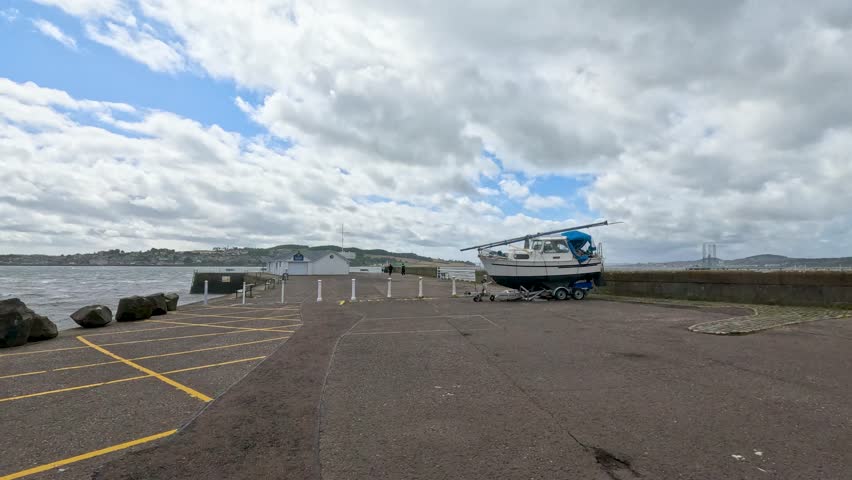 A motor sailer boat is moved on a trailer across an empty coastal parking lot under partly cloudy daylight, with steady wide-angle camera framing