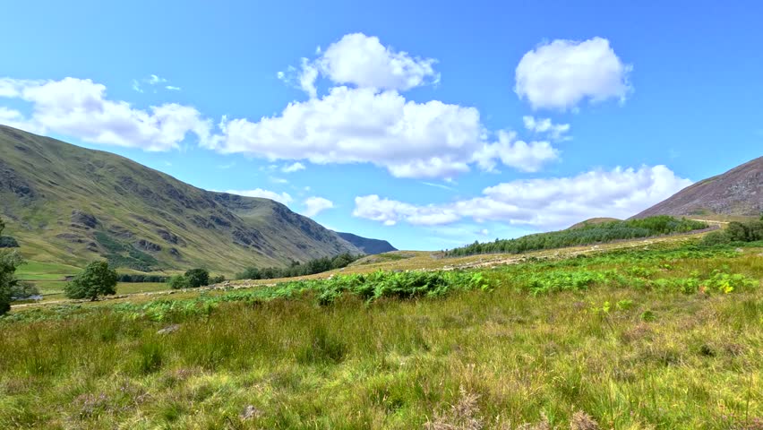 Camera moves steadily along a rugged dirt path through grassy hills under bright daylight, revealing scenic Scottish landscape with blue sky and scattered clouds