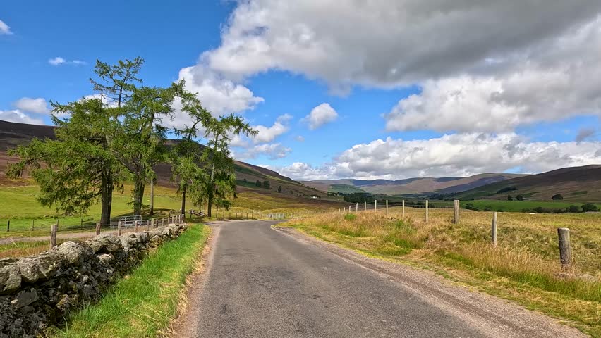 A vehicle travels down a winding country road through scenic Scottish hills under partly cloudy skies, with dynamic forward camera movement and natural daylight