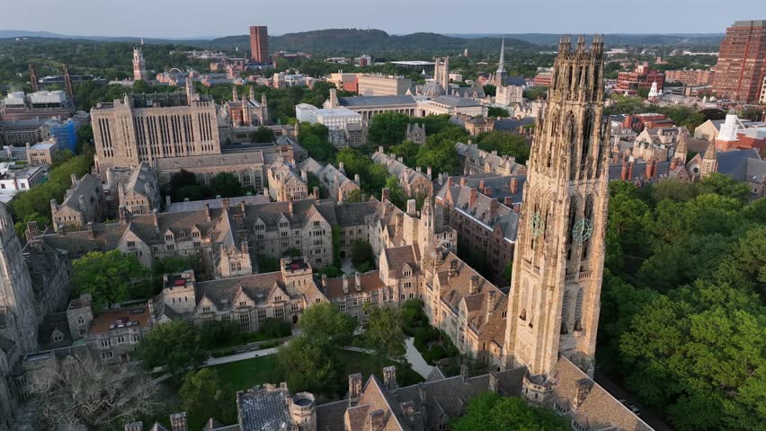 Old campus courtyard with harkness tower in New Haven, Connecticut. Aerial top down rising shot. Sunset time in well-kept park of high school university. Ancient buildings and houses in town.
