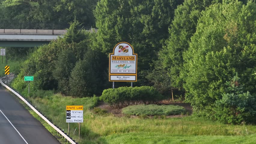 Aerial zoom shot of welcome to Maryland road sign on highway. Truck on bridge crossing. Green trees in summer. Border between American states.