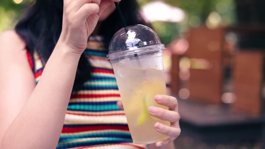 A Asian girl is outdoors, feeling happy and content while enjoying a lemonade drink in the sun. Woman stirring a delicious lemonade at outdoor cafe.