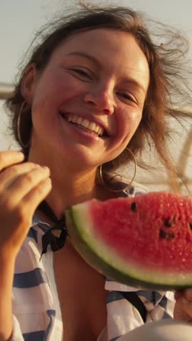 A cheerful woman holds a slice of watermelon, savoring the refreshing taste as golden sunset colors fill the sky during a warm summer evening