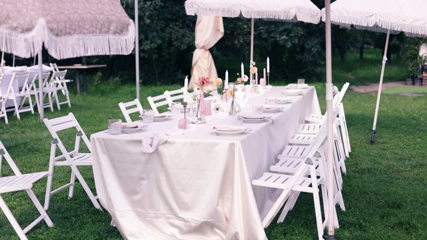 Table neatly set up outside for guests during a wedding reception on a sunny warm day. White wooden chairs set up for wedding ceremony outside house.
