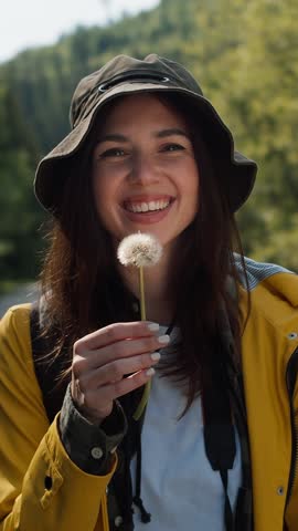A cheerful woman wearing a yellow raincoat holds a fluffy dandelion, smiling brightly as she embraces the beauty of a sunny day in the great outdoors