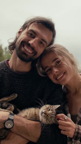 A cheerful couple enjoys a cozy moment outdoors, cradling their beloved cat, surrounded by the beauty of nature and the warmth of companionship