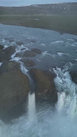 Aerial view of white water cascading down a steep waterfall over dark rocks, creating a misty and dynamic scene, pingeyjarsveit, Iceland.