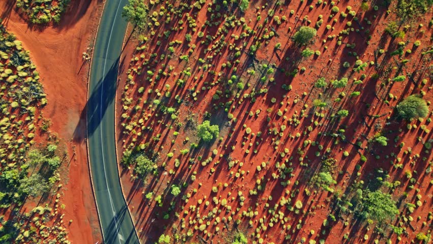 Aerial view of a winding asphalt road cutting through the vibrant red earth and sparse greenery of the Australian outback, Ayers Rock, Northern Territory, Australia.