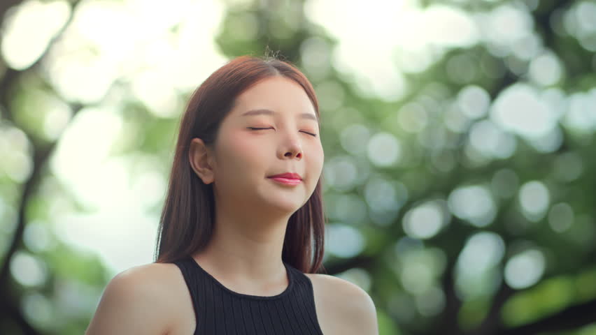 Peaceful asian woman practicing meditation and deep breathing in park for mindfulness and spiritual wellness, embracing fresh air and nature for healing energy balance with closed eyes and calm aura