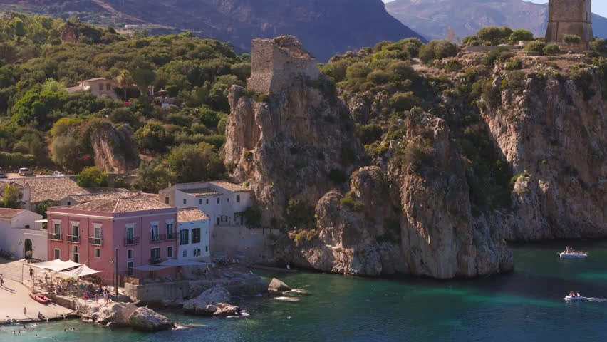 Aerial drone shot lowering down revealing a resort and private beach with huge rocks at Tonnara di Scopello, Sicily, Italy, showcasing the coastline during Italian summer