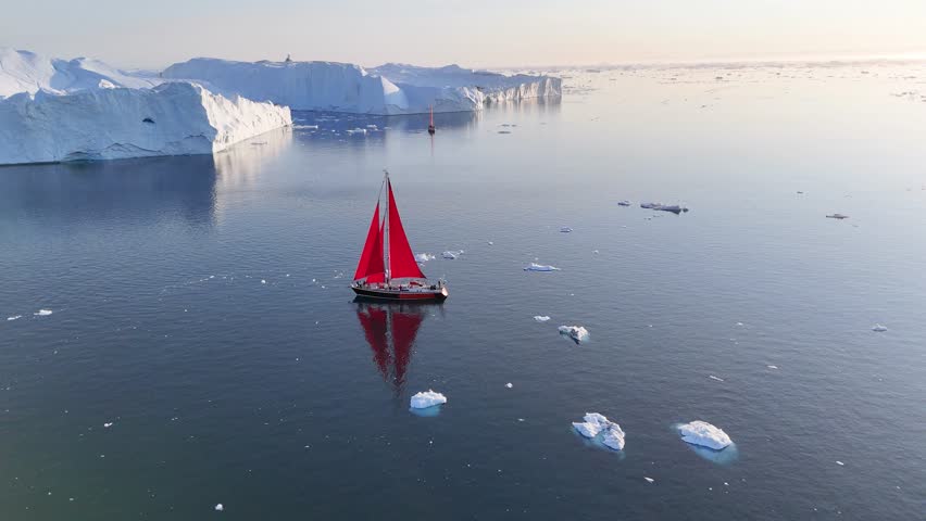 Little red sailboat cruising among floating icebergs in Disko Bay glacier during midnight sun season of polar summer. Ilulissat, Greenland. Global warming and melting glaciers.