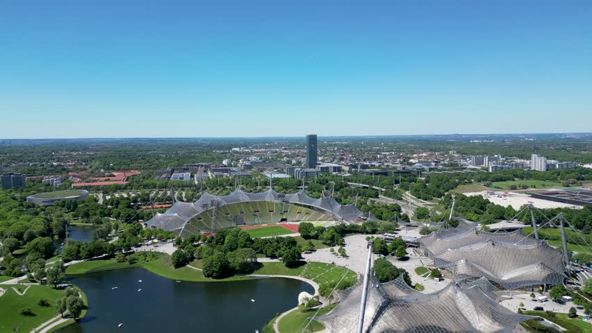 Aerial view of the Olympiapark, stadium, lake and surrounding lush greenery under a clear blue sky, creating a vibrant contrast of colors, Munchen, Bayern, Germany.
