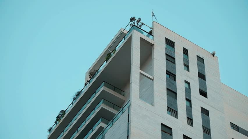 A part of a residential building, in a low angle telephoto view, where a single Israeli flag is hanged out from the top of the apartment building, flapping lightly with the wind. Ramat Gan, Israel.